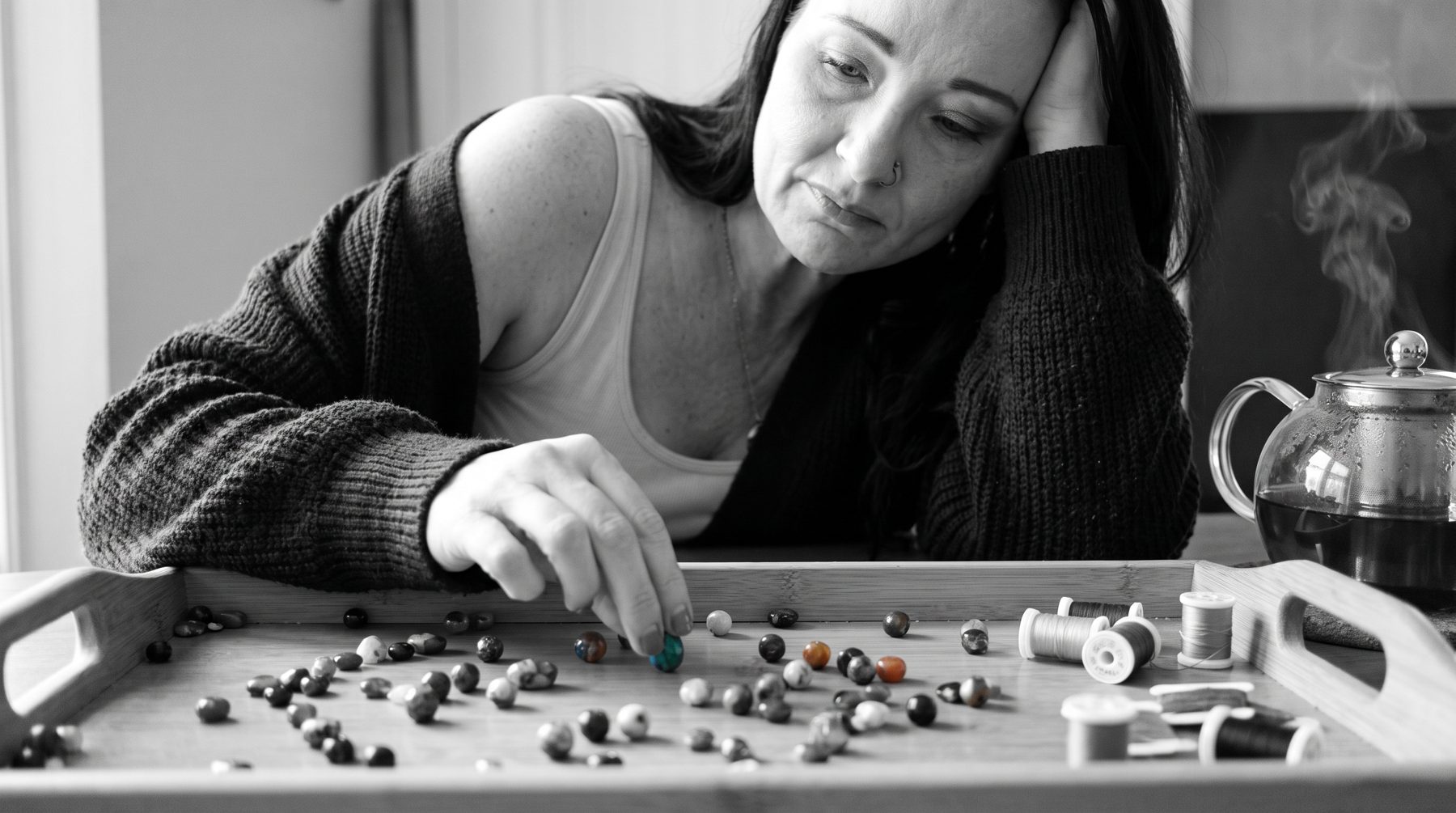 Tiara at the kitchen table with steaming tea, sorting a tray of beads and thread — one bead turquoise in a black-and-white scene.