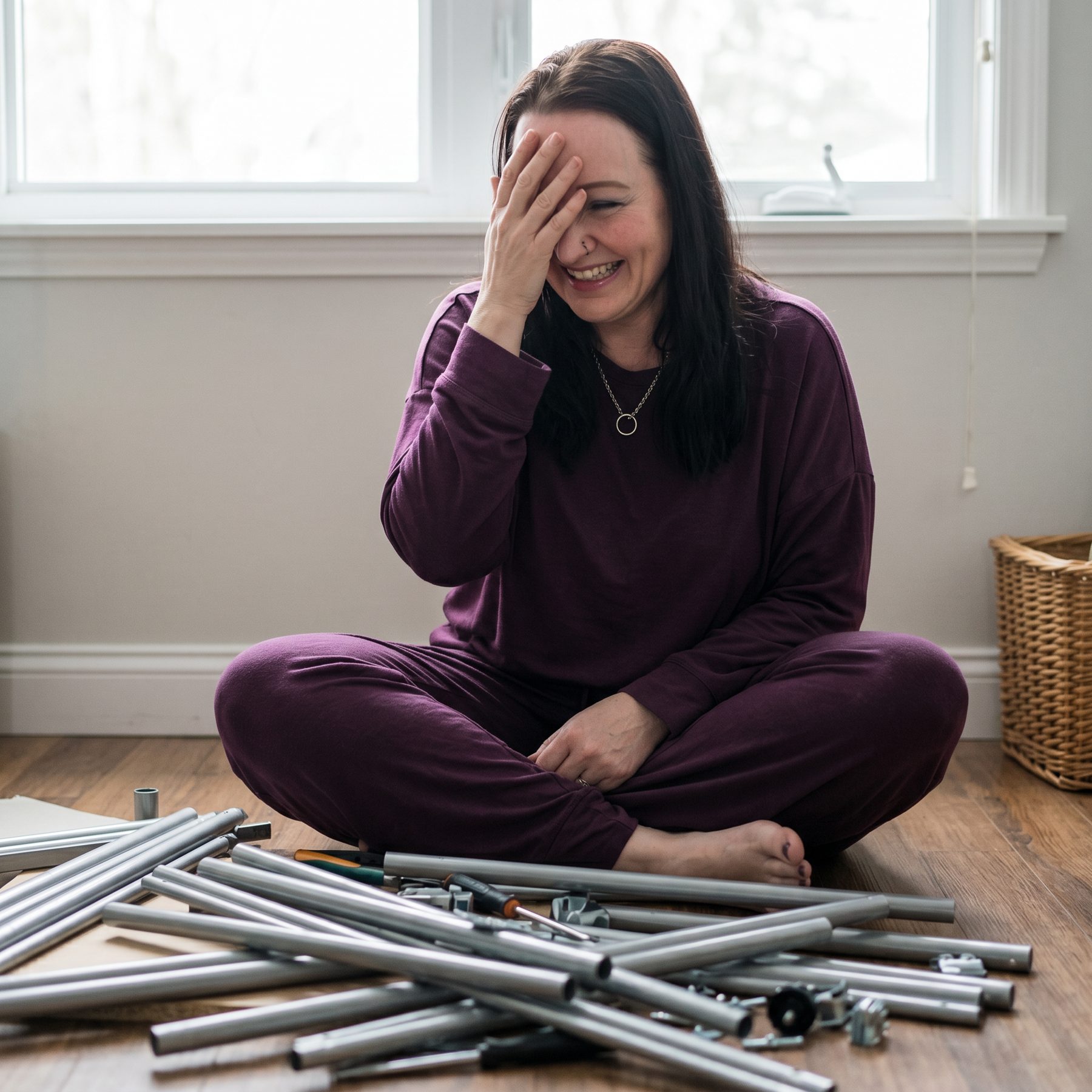 Tiara cross-legged on the floor laughing behind her hand at a pile of unassembled furniture poles.